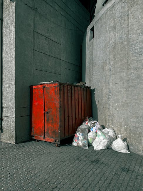 An outdoor urban scene showing a red metal skip with a weathered and rusted surface positioned against a dark green wall and a light grey textured wall. The skip is large, rectangular, and placed on a paved area with a pattern of small, dark paving stones. Next to the skip, there are several white and transparent plastic bags filled with mixed waste, including visible plastic bottles and other debris. The setting appears to be a narrow alley or side street, with the skip used for private or alternative waste disposal, possibly managed by a waste removal service like Rubbish Removal Harringay. The environment is illuminated evenly, with natural daylight highlighting the textures and colors of the skip, walls, and rubbish, emphasizing the practical nature of on-site rubbish collection in an urban context.