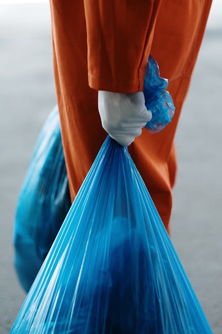Close-up view of a person wearing orange protective overalls and white gloves, holding a large blue plastic waste bag filled with rubbish. The individual's hand is gripping the top of the bag, with the bag's material appearing shiny and slightly translucent. The background is blurred but suggests an outdoor environment, possibly a waste collection or clearance site, with neutral tones. The scene emphasizes the act of handling waste material, consistent with rubbish removal services like those provided by Rubbish Removal Harringay. The focus on protective clothing and waste bags reflects on-site clearance or private disposal efforts, supporting the context of alternative rubbish collection methods. The lighting is neutral and natural, highlighting the textures of the plastic bag and the clothing's fabric, creating a clear, professional visual relevant to waste management and rubbish removal activities.
