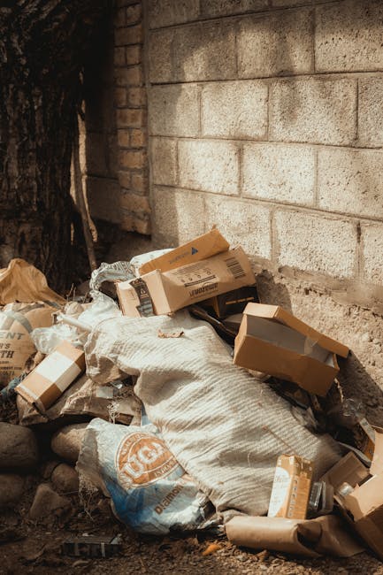 An outdoor urban scene showing a red metal skip with a weathered and rusted surface positioned against a dark green wall and a light grey textured wall. The skip is large, rectangular, and placed on a paved area with a pattern of small, dark paving stones. Next to the skip, there are several white and transparent plastic bags filled with mixed waste, including visible plastic bottles and other debris. The setting appears to be a narrow alley or side street, with the skip used for private or alternative waste disposal, possibly managed by a waste removal service like Rubbish Removal Harringay. The environment is illuminated evenly, with natural daylight highlighting the textures and colors of the skip, walls, and rubbish, emphasizing the practical nature of on-site rubbish collection in an urban context.
