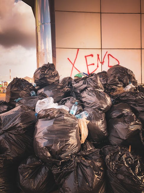 A large pile of black rubbish bags, some with visible torn sections, stacked against a modern tiled wall with a reflective metal column on the left side. The bags contain various waste items, including partially visible plastic bottles and packaging materials, with some plastic bottles seen protruding from the bags. Behind the pile, the wall features red graffiti text reading 'XEND' in spray paint. The environmental setting appears to be an urban outdoor area, possibly a designated waste collection point or an unmanaged rubbish accumulation site. The lighting suggests late afternoon or early evening, casting a warm hue over the scene. This scene highlights issues related to private waste disposal and the importance of proper rubbish removal services, such as those provided by Rubbish Removal Harringay, especially when access for collection is obstructed or when independent disposal methods are used, contributing to local waste management challenges.