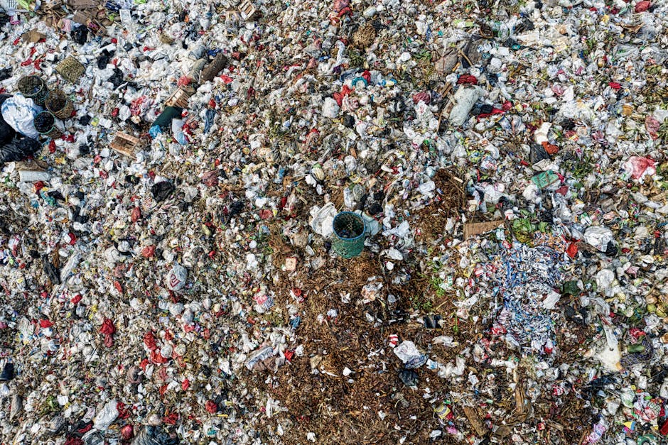 An extensive landfill area filled with a large quantity of mixed waste, predominantly composed of crumpled paper, plastic wrappers, and various debris. The waste appears to be a combination of household rubbish and discarded packaging, with some items partially buried under others. There are scattered pieces of cardboard, plastic bottles, and foil packaging, creating a dense and chaotic surface. In the central section, a small blue plastic bucket is partially buried amidst the refuse, surrounded by soil and compacted waste materials. The background shows more piles of rubbish extending into the distance, with no visible vegetation or natural elements. The lighting indicates an overcast day, casting a diffuse light over the scene, emphasizing the varied textures and colours of the waste. This detailed view illustrates the type of clutter that could hinder access for waste collection services, highlighting the importance of professional rubbish removal and on-site clearance by companies like Rubbish Removal Harringay, especially for private disposal or alternative waste handling scenarios.