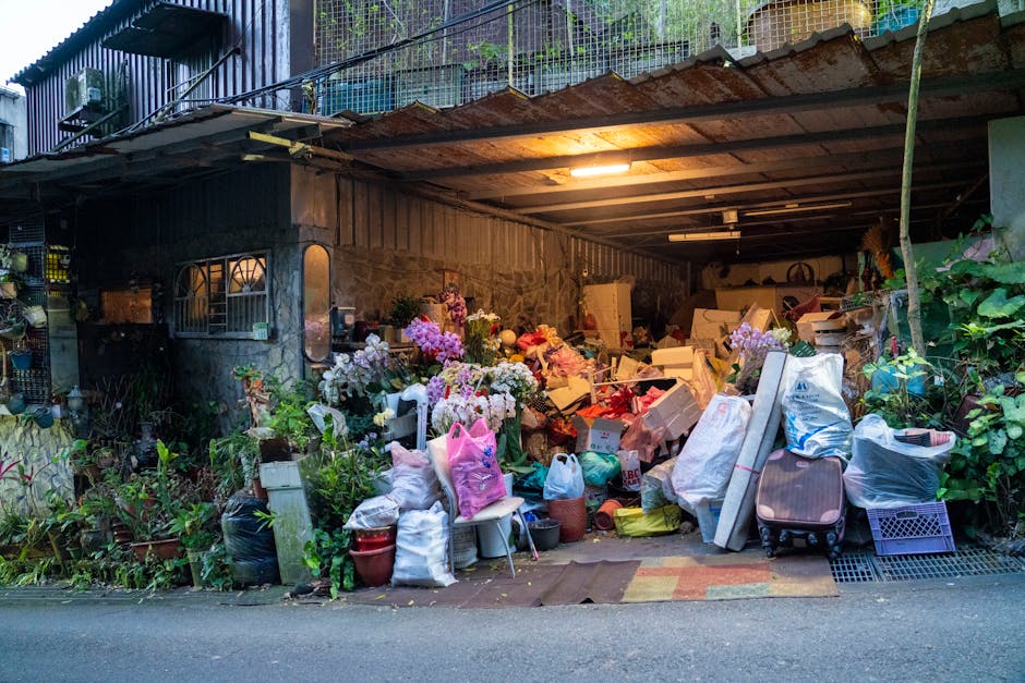 An outdoor storage area adjacent to a building, featuring a variety of discarded items including cardboard boxes, plastic bags, and household objects piled loosely on the ground and against the wall. The space is partially covered by a rusted metal roof supported by metal beams, with an exposed wooden underside. The background includes a stone wall and a section of metal fencing, with some greenery growing along the edges of the pile. Several potted plants and flowers, some in full bloom, are placed among the rubbish, contrasting with the clutter. The environment is illuminated by a single internal light source, casting a warm glow over the scene. The clutter appears temporary, suggesting a collection of waste or items awaiting removal, fitting within the context of private rubbish clearance services typically offered by companies like Rubbish Removal Harringay, especially for hoarded or overloaded storage spaces.