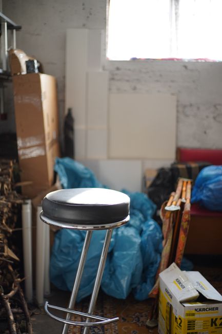 The image shows a cluttered indoor space with various discarded items and waste materials. In the foreground, there is a black and silver stool with a round cushioned seat and metal legs, positioned near the bottom edge. Behind the stool, several large blue plastic garbage bags filled with unknown rubbish are piled against a white brick wall, some of which are partially open revealing contents. To the left, a tall wooden shelving unit holds miscellaneous objects, including a cardboard box and smaller items stacked on shelves. Leaning against the wall are white panels or boards, and a partially visible window above allows natural light to illuminate the scene, giving a neutral and pragmatic appearance. On the right, a cardboard box labeled 'KARCHER' containing cleaning equipment is situated near a piece of furniture with a red cushion, possibly a sofa or bed frame. The environment suggests an on-site clearance or private waste collection process that a company like Rubbish Removal Harringay might handle as part of their rubbish removal services in Harringay, capturing a typical cluttered storage or hoarded room awaiting clearing or disposal, emphasizing the need for professional rubbish removal solutions. The overall scene is characterized by disorder and a mixture of materials indicative of household or renovation waste within an interior setting.