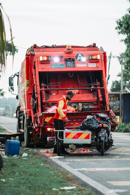 A street scene showing a worker in a red and yellow uniform operating a large red waste collection truck parked at the roadside. The back of the truck is open, revealing mechanical components and a collection chamber, with the worker engaging in rubbish collection or disposal activities. Nearby, a motorcycle with a black plastic bag secured on its seat is present, along with a small metal trolley or bin positioned close to the worker. The background features a partly cloudy sky, trees, and a parked signboard on the right side of the image. The environment appears to be an urban or suburban street, with a pavement and some scattered litter on the ground. The scene illustrates a private waste collection activity, aligning with alternative rubbish removal services such as those offered by Rubbish Removal Harringay, emphasizing on-site or independent rubbish disposal tools and equipment being used in a typical roadside setting.