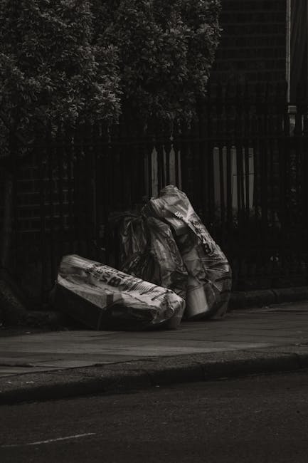 On the pavement beside a black metal fence, three large black plastic rubbish bags filled with waste are stacked together. The bags appear to be heavy and are slightly crumpled, with some visible text and branding printed on their surfaces. Behind the fence, dense foliage of a tree with dark, leafy branches is partially visible, casting shadows on the scene. The setting appears to be outdoors in an urban or residential area, with the bags likely awaiting collection or disposal. The image’s lighting is subdued, suggesting either overcast weather or heading towards evening. This scene exemplifies a typical private rubbish removal situation, where waste is temporarily stored on the street for an alternative collection service to manage, aligning with the principles of independent waste disposal often handled by companies like Rubbish Removal Harringay.