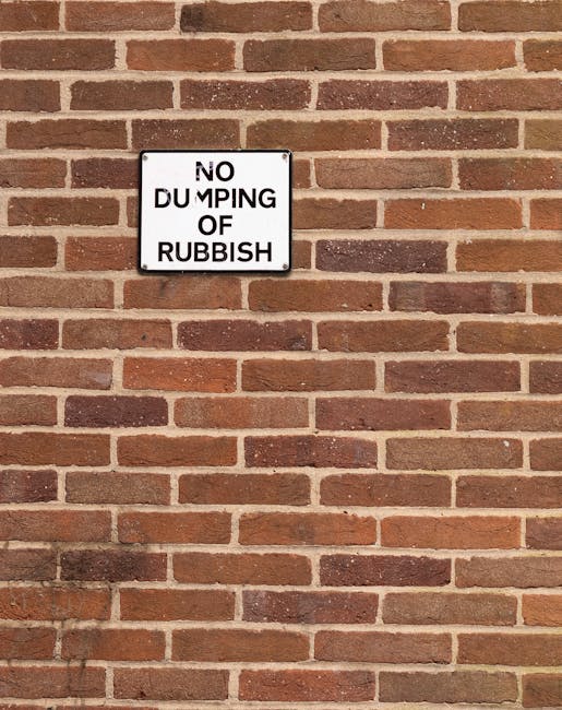 A rectangular white metal sign mounted on a red brick wall, displaying the message 'NO DUMPING OF RUBBISH' in black capital letters, with 'DUMPING' partially obscured by wear or damage. The brick wall has a traditional running bond pattern with reddish-brown bricks and light gray mortar joints. The sign is positioned near the upper left area of the image, and the lighting appears natural, casting slight shadows on the wall. The setting suggests a maintained outdoor environment, typical of areas where rubbish removal services or regulations are relevant, and highlights the importance of adhering to proper waste disposal practices, potentially supported by local authorities or private rubbish collection services such as Rubbish Removal Harringay.