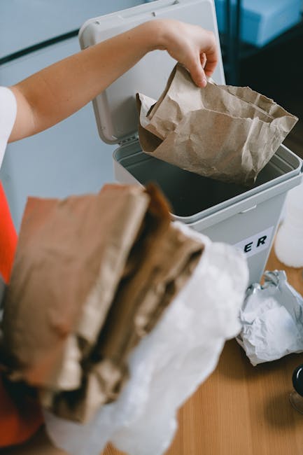 A person is seen disposing of paper waste into a grey rubbish bin with a partially open lid, situated indoors on a wooden surface. The person's arm is extended, holding a crumpled brown paper bag, which is being placed inside the bin. In the foreground, there are additional crumpled brown and white paper bags, some partially obscured. The environment appears to be a domestic setting, with a focus on waste disposal, relevant to private rubbish collection or on-site clearance services offered by Rubbish Removal Harringay. The bin is positioned against a light-colored wall, with natural or ambient indoor lighting illuminating the scene clearly, emphasizing the textures of the paper and the smooth surface of the bin. This image visually relates to the topic of effective household rubbish management and alternative waste handling methods, illustrating typical manual waste segregation and disposal practices.