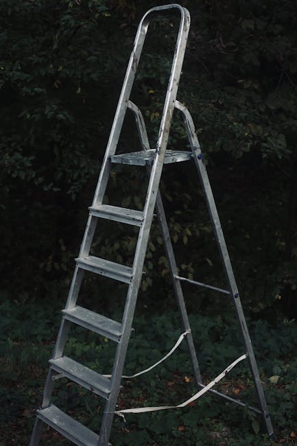 A tall, silver aluminium step ladder with four steps and a top platform, positioned outdoors against a backdrop of dense, dark green foliage and shrubbery. The ladder's rungs have a textured surface for grip, and a safety strap is loosely secured around the lower part of the ladder, indicating it may be used for maintenance or access purposes. The environment appears to be a garden or backyard area with natural daylight illuminating the scene, highlighting the ladder's metallic finish and the surrounding foliage. This scene relates to private waste handling or on-site clearance, which Rubbish Removal Harringay's services may support through alternative rubbish collection options. The overall setting is neat, with no visible clutter or debris around the ladder, emphasizing a straightforward outdoor scene suitable for rubbish or equipment removal preparations.