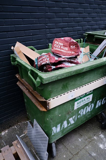 A large green wheeled skip placed on a paved outdoor surface next to a dark brick wall, filled with red and black plastic bags containing garden or construction waste, with some bags displaying text about flex and garden supplies. The skip is situated on a metal stand and appears to be part of a private waste collection service, possibly used for onsite clearance or alternative rubbish removal. Nearby, a white brick filter indicating the service name and contact details is visible, with a small section of a corrugated metal sheet partially leaning against the skip. The scene depicts a typical outdoor setting for waste disposal, associated with independent waste management or rubbish removal in Harringay, aligned with non-Council rubbish collection practices.