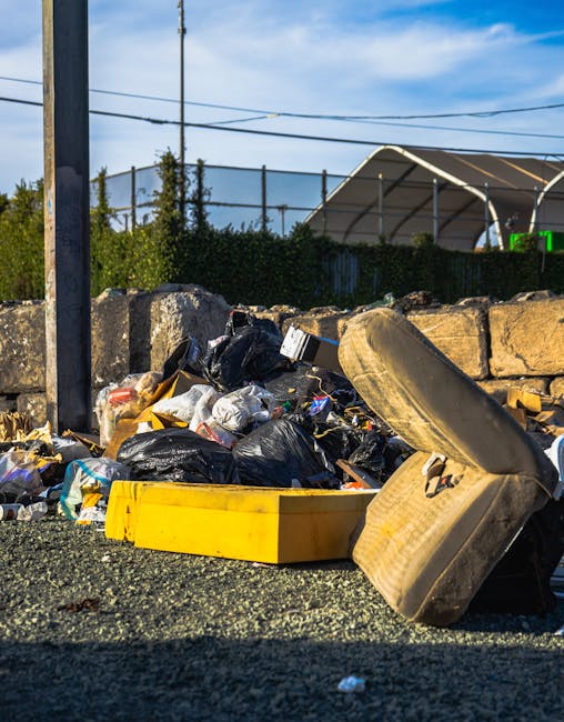 A large green wheeled skip placed on a paved outdoor surface next to a dark brick wall, filled with red and black plastic bags containing garden or construction waste, with some bags displaying text about flex and garden supplies. The skip is situated on a metal stand and appears to be part of a private waste collection service, possibly used for onsite clearance or alternative rubbish removal. Nearby, a white brick filter indicating the service name and contact details is visible, with a small section of a corrugated metal sheet partially leaning against the skip. The scene depicts a typical outdoor setting for waste disposal, associated with independent waste management or rubbish removal in Harringay, aligned with non-Council rubbish collection practices.
