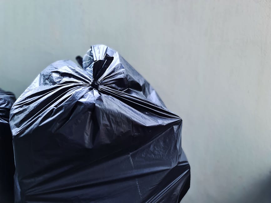 A tightly tied black plastic rubbish bag with a glossy surface, placed in front of a plain, light-colored wall. The bag appears full and slightly bulging, with some creases and folds visible on its surface. In the background, other black rubbish bags are stacked or leaning against each other, indicating a collection of waste awaiting disposal. The setting suggests an outdoor or indoor space designated for rubbish storage, such as a driveway, alleyway, or waste collection area. The lighting is neutral, casting soft shadows on the bags and highlighting the reflective quality of the plastic material. This scene aligns with waste management practices and could relate to private rubbish collection services provided by companies like Rubbish Removal Harringay, especially in contexts where independent or supplementary waste disposal is performed outside of local authority collection services.