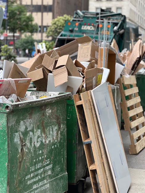 A large green wheeled dumpster contains a significant amount of waste, including brown corrugated cardboard boxes, some of which are flattened and others partially assembled, with visible packing tape and labels. The boxes are stacked haphazardly, overflowing and leaning against the sides of the container. In front of the dumpster, there are wooden pallets, some with metal or plastic components, positioned vertically or leaning against the side. The scene appears to be outdoors on an urban street, with a large waste collection vehicle or skip visible in the background, partially obscured by the accumulated rubbish. The surrounding environment includes blurred city buildings and trees, suggesting a busy area for waste collection or disposal. The lighting indicates it is daytime, with natural sunlight illuminating the scene, highlighting the textures of the cardboard, wood, and metal surfaces. The image relates to private waste handling and rubbish removal services, such as those offered by Rubbish Removal Harringay, emphasizing the process of on-site clearance or alternate rubbish collection methods used in urban environments.
